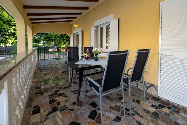 Un patio techado al aire libre con suelo de mosaico, una mesa y sillas, y vista a la vegetación a través de la puerta abierta.