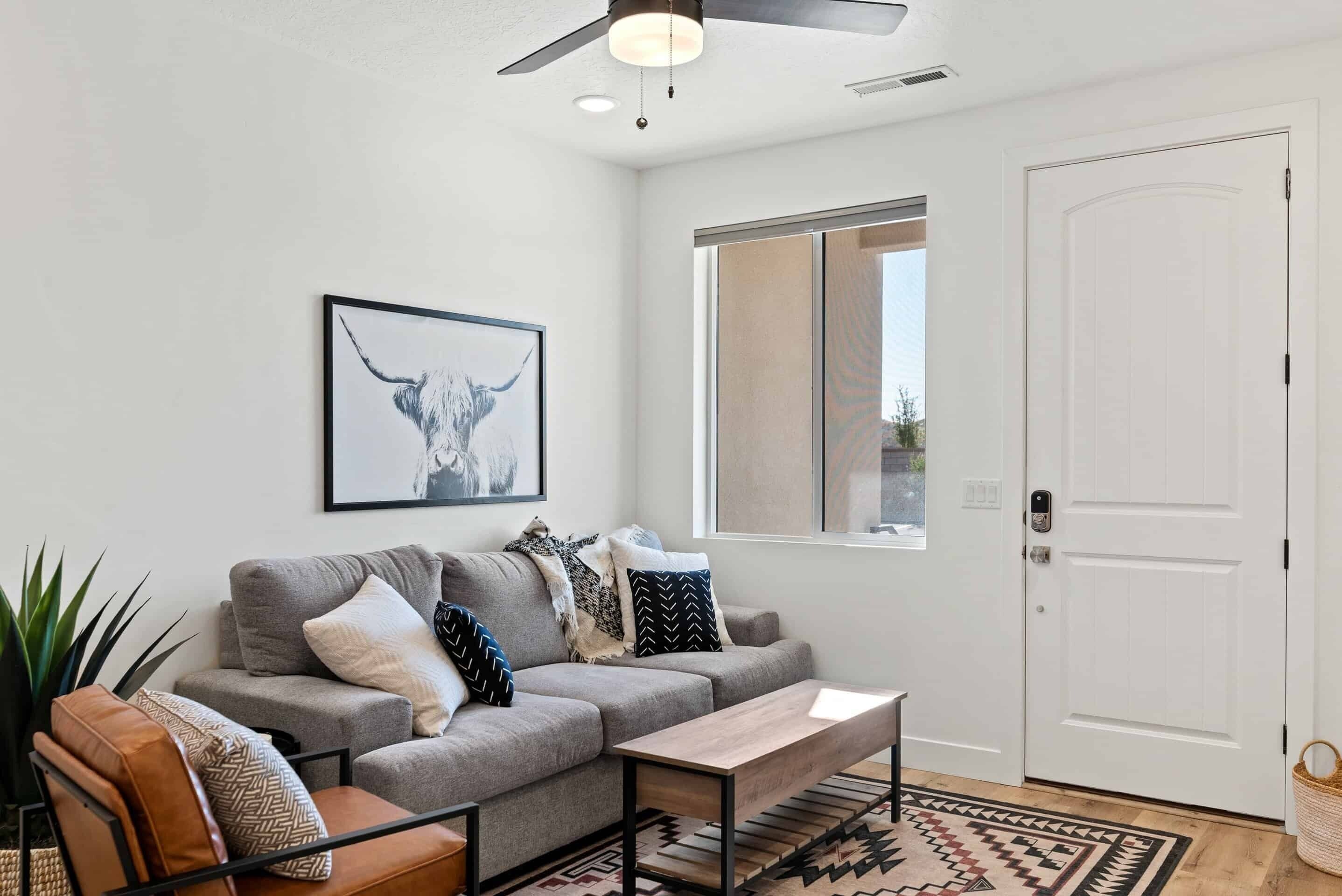 Ein Wohnzimmer mit grauem Sofa, Couchtisch, Deckenventilator und großem Fenster.