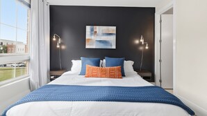 Een opgemaakt bed met een blauwe deken en twee kussens, een kunstwerk aan de muur, en twee bedlampjes.