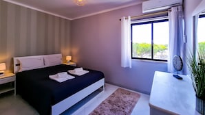 A modern bedroom with a bed, bedside lamps, a window with curtains, an air conditioning unit, and a potted plant. - FLH Vilamoura Balcony with Pool (Vilamoura)