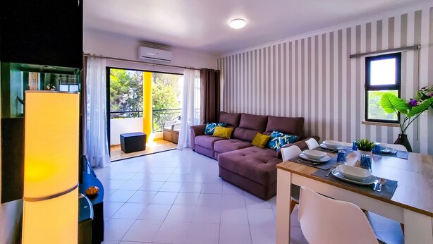 A modern living room with a sofa, dining table, and chairs. There is a balcony with a view of trees and a window with curtains. - FLH Vilamoura Balcony with Pool (Vilamoura)