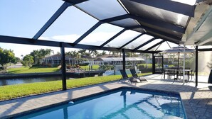 A swimming pool with a glass enclosure, a patio area with chairs, and a view of a residential area with houses and a dock. -  Private enclosed heated pool with Gulf access. Panoramic Lenai (Cape Coral)