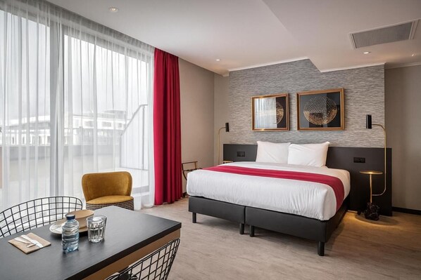 A hotel room with a bed, a desk with a chair, a window with sheer curtains, and a red curtain.