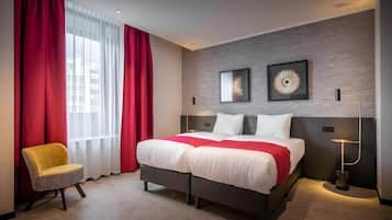 Ett hotellrum med en säng, röda gardiner, en stol och två konstverk på väggen.