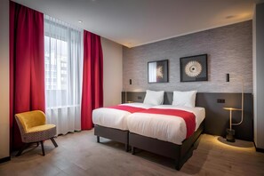 A hotel room with a bed, red curtains, a chair, and two wall art pieces. - Amory Hotel (Antwerp)