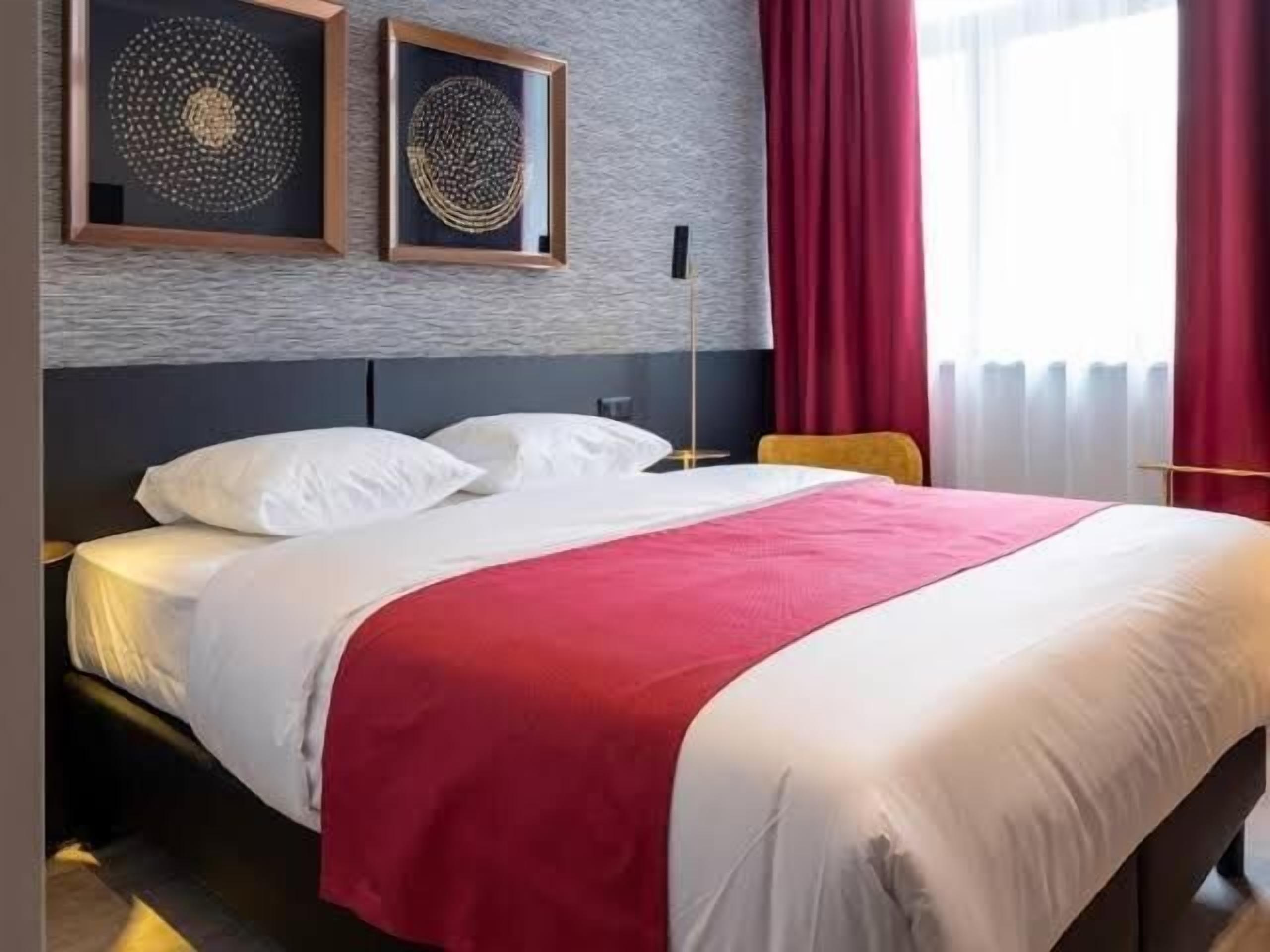 A hotel room with a bed, two framed artworks on the wall, a red curtain, and a window with white curtains.