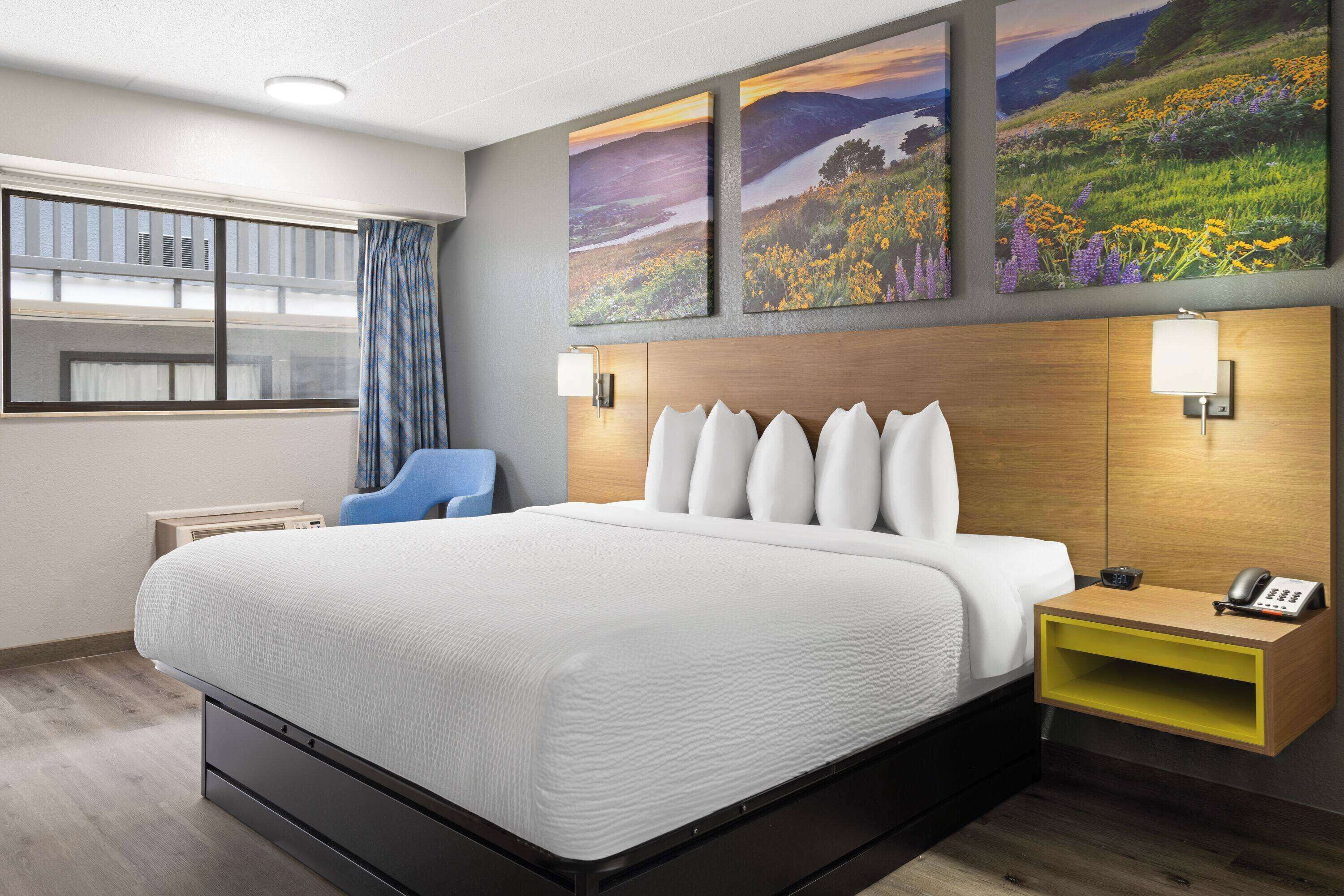 Una habitación de hotel con una cama grande, una mesita de noche con lámpara, una silla y una obra de arte en la pared.