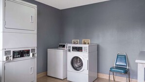 A laundry room with a front-loading washer and dryer, a blue chair, and a small table with framed pictures. - Days Inn by Wyndham Columbus/Worthington (Columbus)