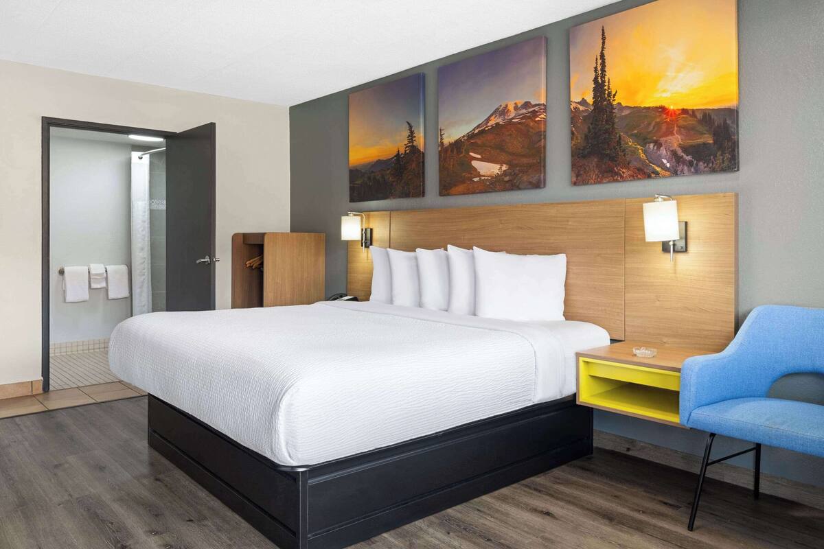 A hotel room with a large bed, a wooden headboard, two wall-mounted lamps, a blue chair, and three paintings of mountains.