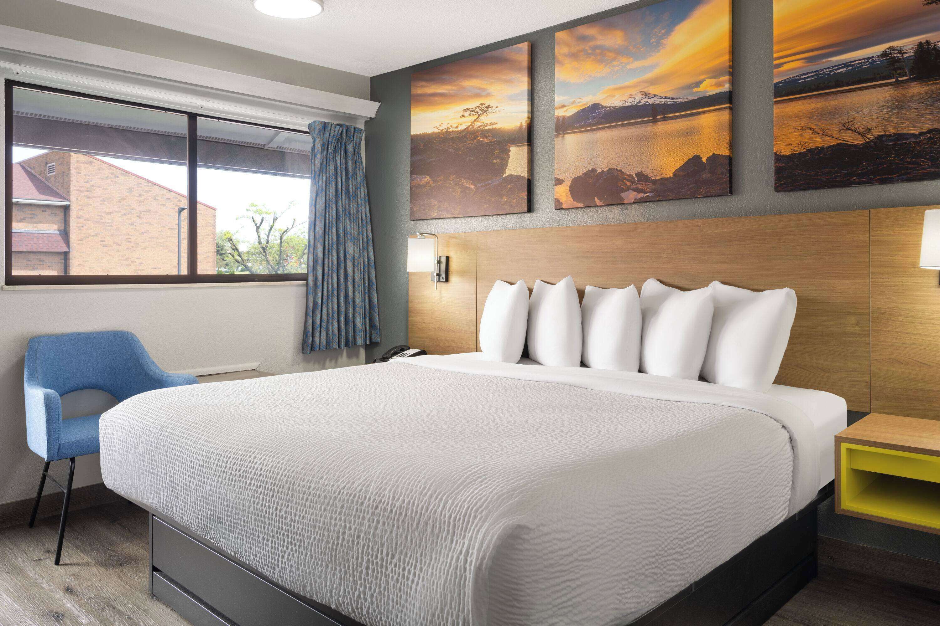 Ett hotellrum med en stor säng, en blå stol, en sänggavel i trä och utsikt över en byggnad genom ett fönster.