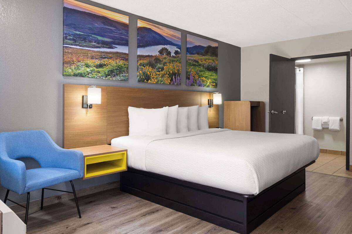 A modern hotel room with a large bed, a blue chair, a wooden nightstand, and wall-mounted lamps.