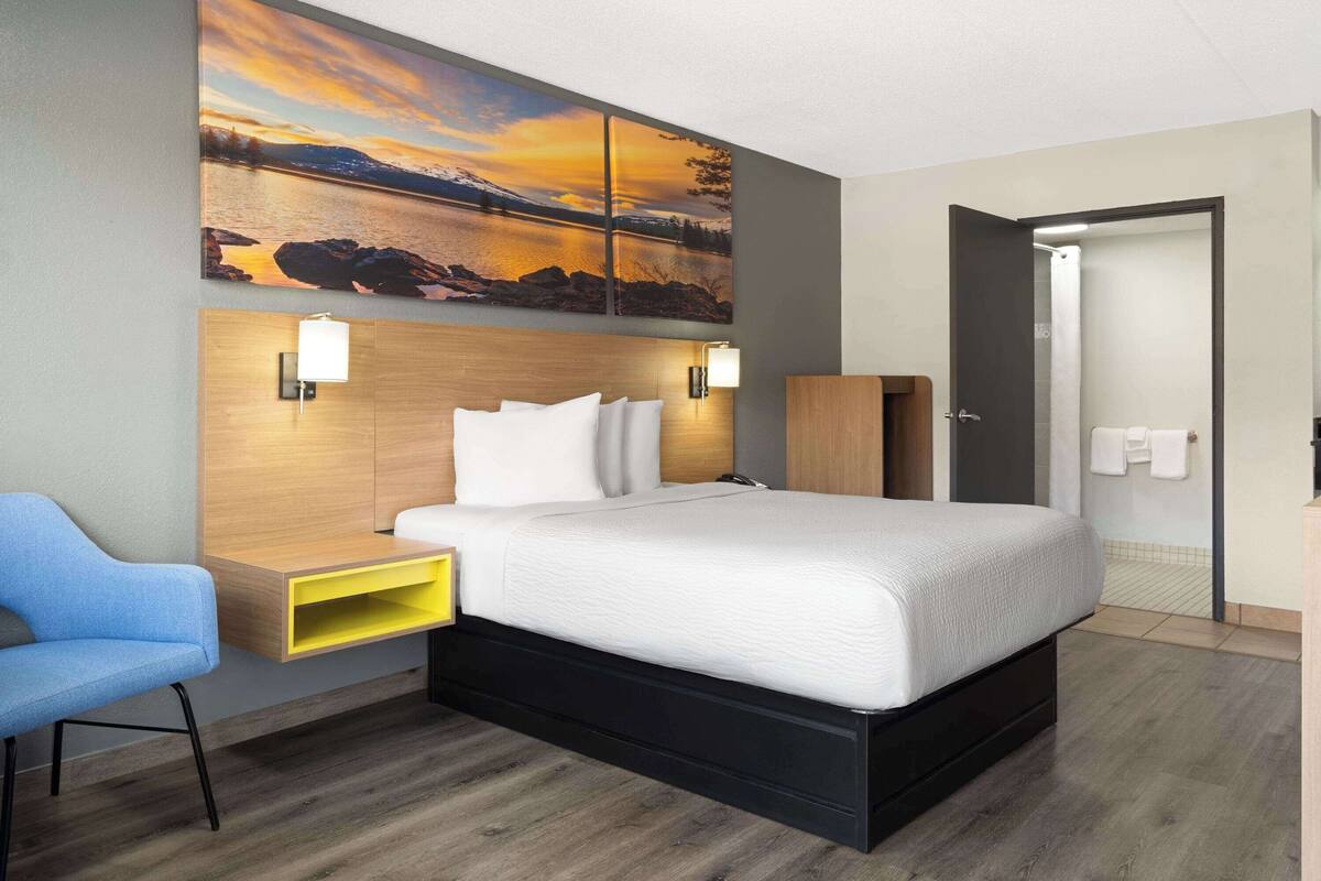 A modern hotel room with a large bed, a nightstand, a desk, a chair, and a painting of a sunset over a lake.