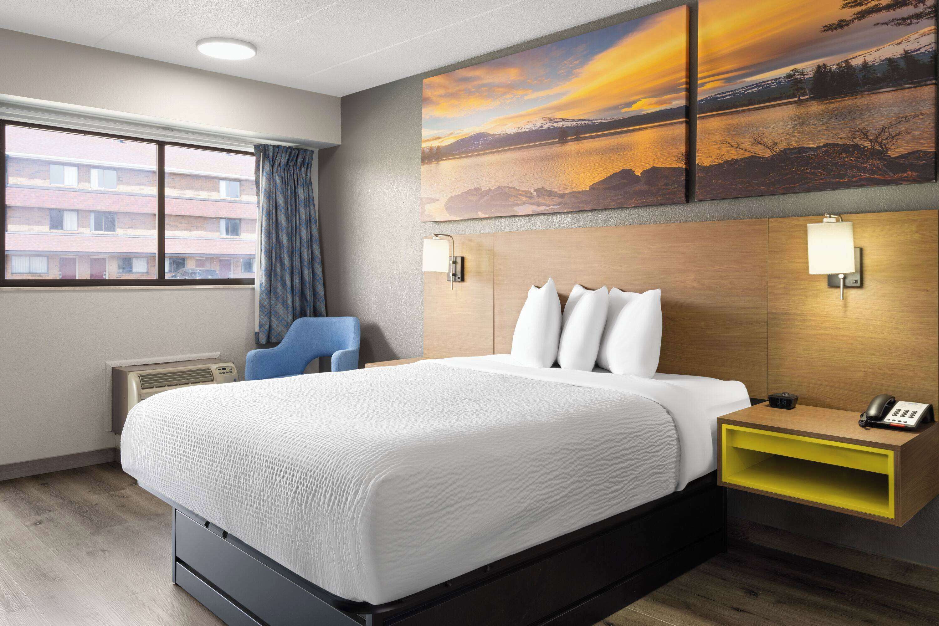 A hotel room with a large bed, a desk with a phone, and a painting of a sunset over a lake.