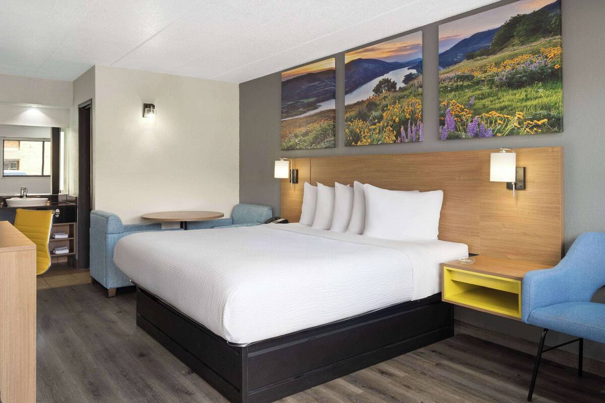 A hotel room with a large bed, a desk, a chair, and a small table. There are three paintings on the wall above the bed.
