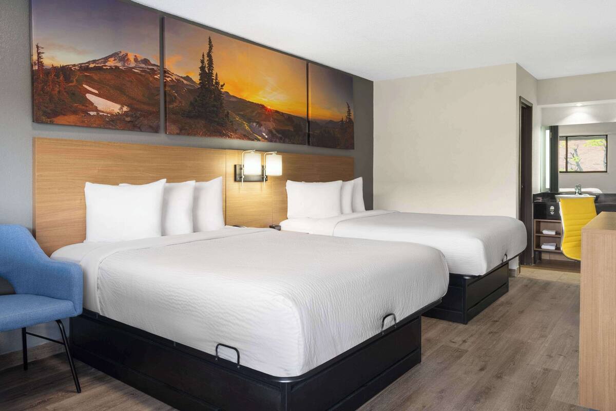 A hotel room with two beds, a wooden headboard, a wall-mounted light, and a painting of a mountain landscape.