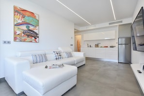 Una sala de estar moderna con un sofá blanco, un cuadro de peces coloridos y una cocina con armarios blancos y un refrigerador de acero inoxidable.