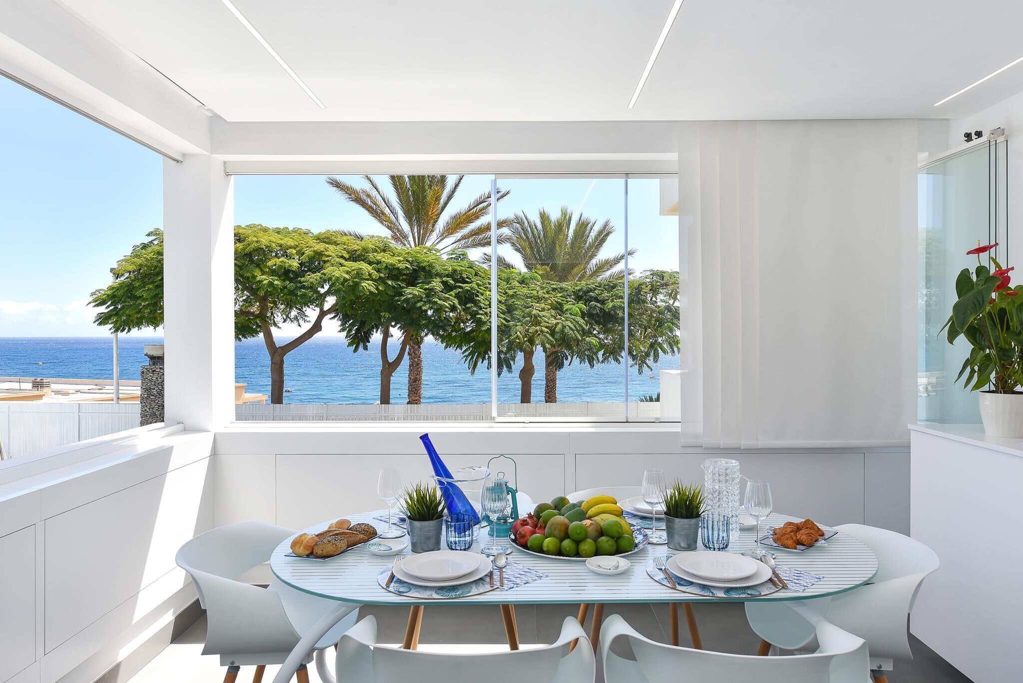 Un espace de restauration moderne avec une table en verre, des chaises blanches et une vue sur la mer et les palmiers.