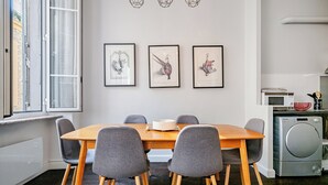 A dining area with a wooden table and grey chairs, a window with curtains, and three framed pictures on the wall. - Stunning apartment in the Gold Square of Nice! (Nice)