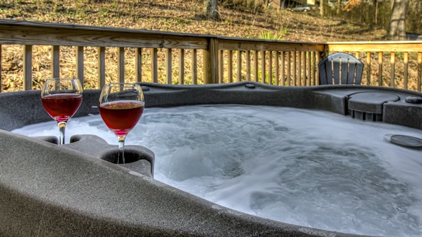 Un bain à remous avec deux verres de vin rouge, entouré d’une terrasse en bois et offrant une vue sur les arbres et les maisons au loin.