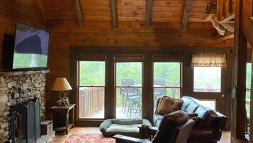 A cozy cabin interior with a stone fireplace, a wooden ceiling, a leather sofa, and a large window overlooking a deck.