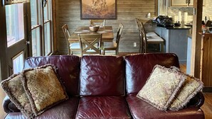 Ein rustikales Blockhäuschen im Inneren, mit Holzbalkendecke, Essbereich mit Tisch und Stühlen sowie einer Ledersofa mit gemusterten Kissen.
