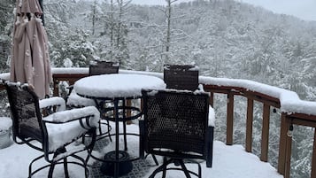 A snow-covered outdoor seating area with a round table and four chairs, surrounded by a wooden deck railing.