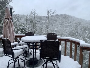A snow-covered outdoor seating area with a round table and four chairs, surrounded by a wooden deck railing. - Private Mountain View cabin on 5 acres, great patio with creek below back patio. (Talking Rock)