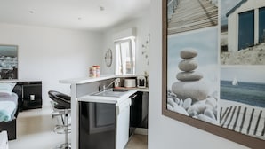 Una habitación de hotel moderna con kitchenette, una cama, zona de estar y una gran obra de arte costera enmarcada.