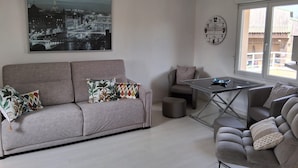 Un moderno salón con un sofá gris, una mesita pequeña y un sillón. En la pared, un cuadro grande de un paisaje urbano en blanco y negro, un reloj redondo y una ventana con vistas a edificios.