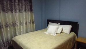 A bed with a patterned bedspread and two pillows, a wooden nightstand, and a curtain with a floral design. - Home away from home (Nyeri)