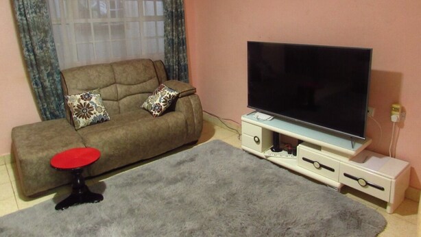 A living room with a sofa, a red stool, a TV on a stand, and a grey rug. - Home away from home (Nyeri)