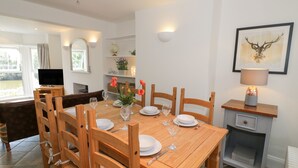 Un coin repas avec une table en bois dressée pour six personnes, entourée de chaises en bois. Un téléviseur est posé sur un meuble à gauche, une étagère avec des objets décoratifs se trouve au-dessus, et une image encadrée orne le mur à droite.