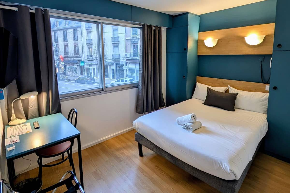 A hotel room with a bed, a desk, and a chair. There is a window with curtains, and a view of buildings outside.