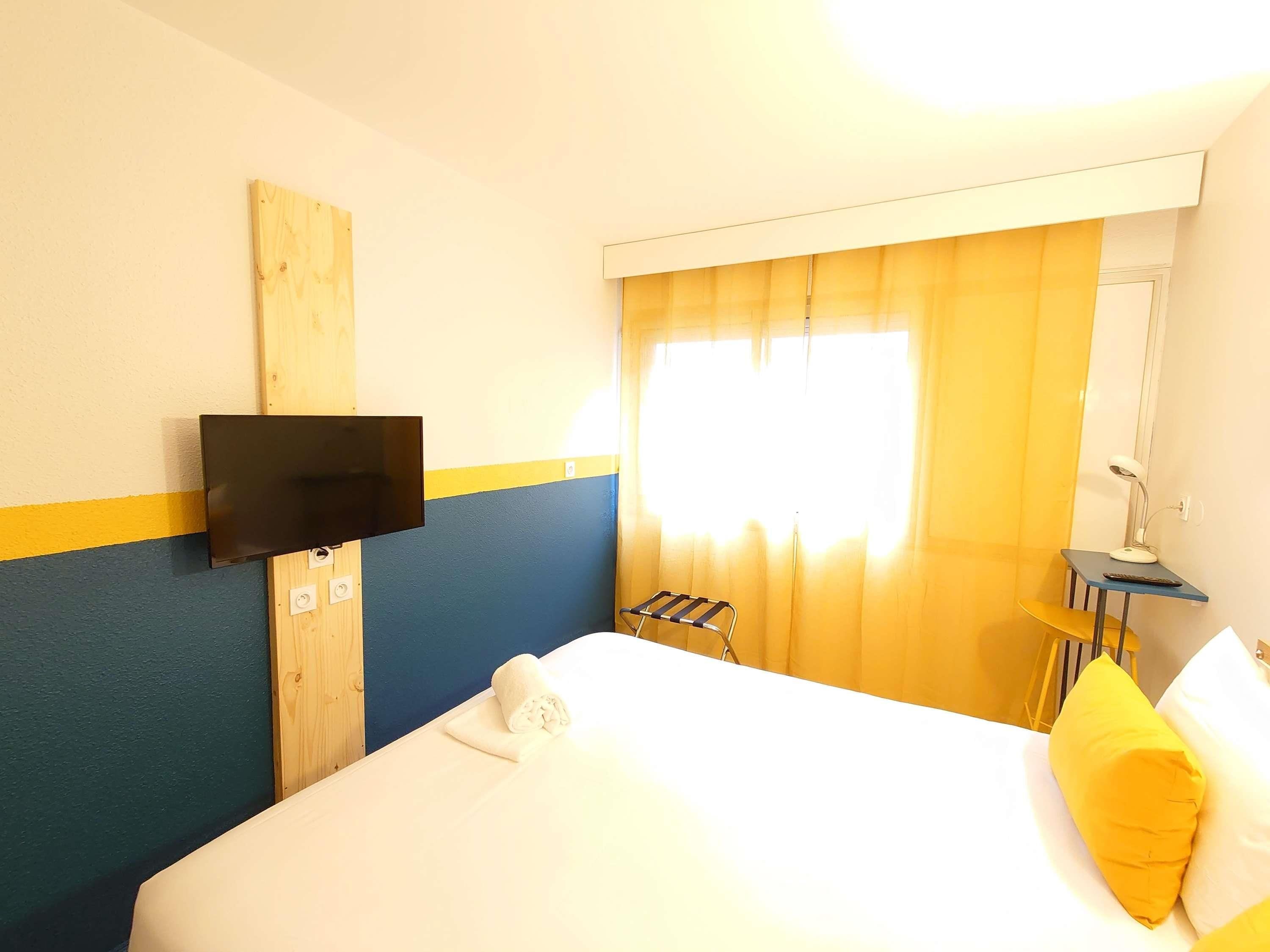 A hotel room with a bed, a TV mounted on the wall, a window with curtains, a desk with a lamp, and a chair.