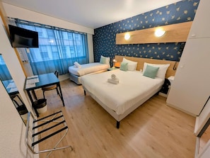 Una habitación de hotel con cama, escritorio, silla, televisor y ventana con cortinas.