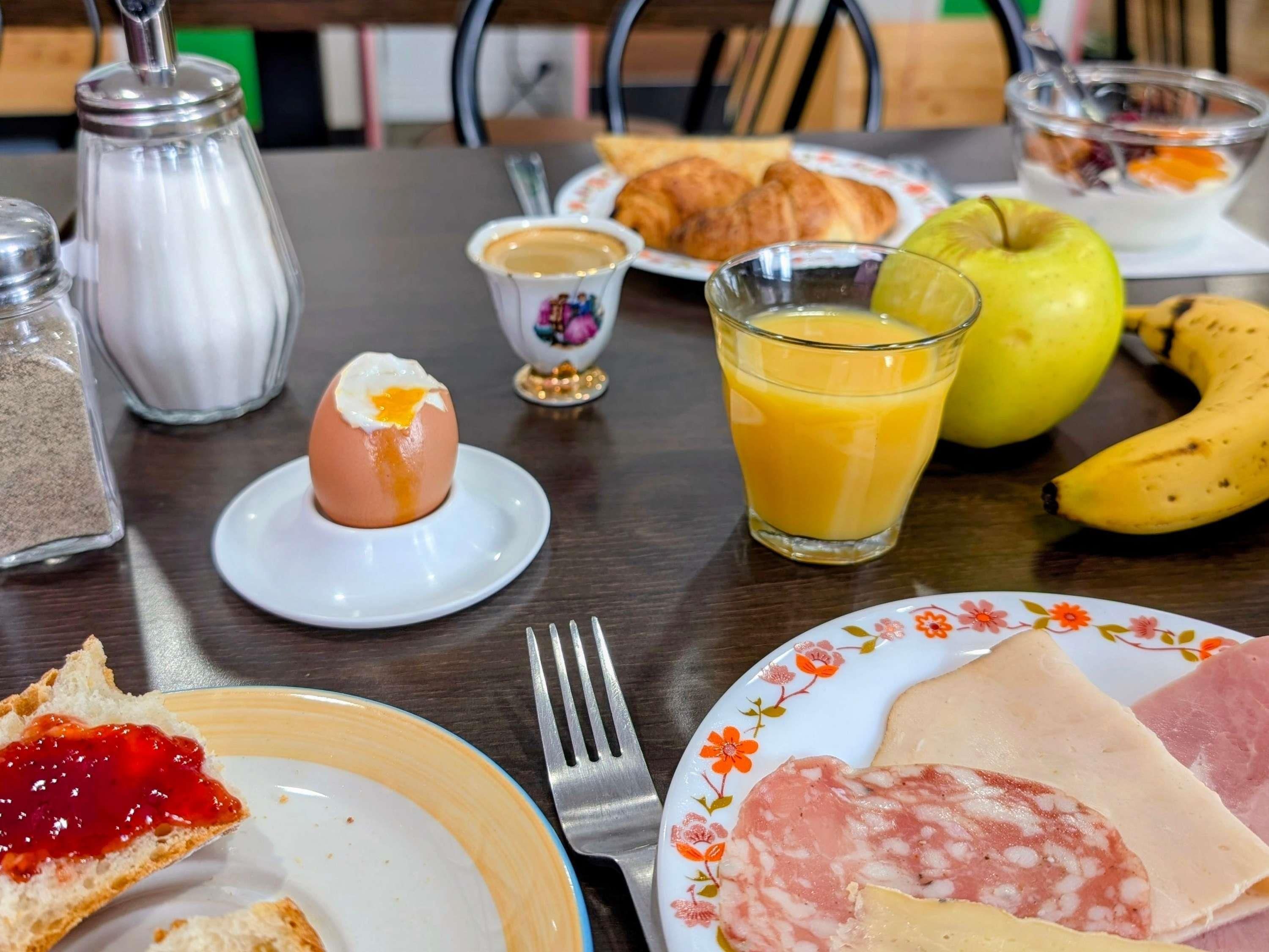 Een ontbijttafel met croissants, een gekookt ei, een glas sinaasappelsap, een banaan, een boterham met ham, en een bordje met jam.