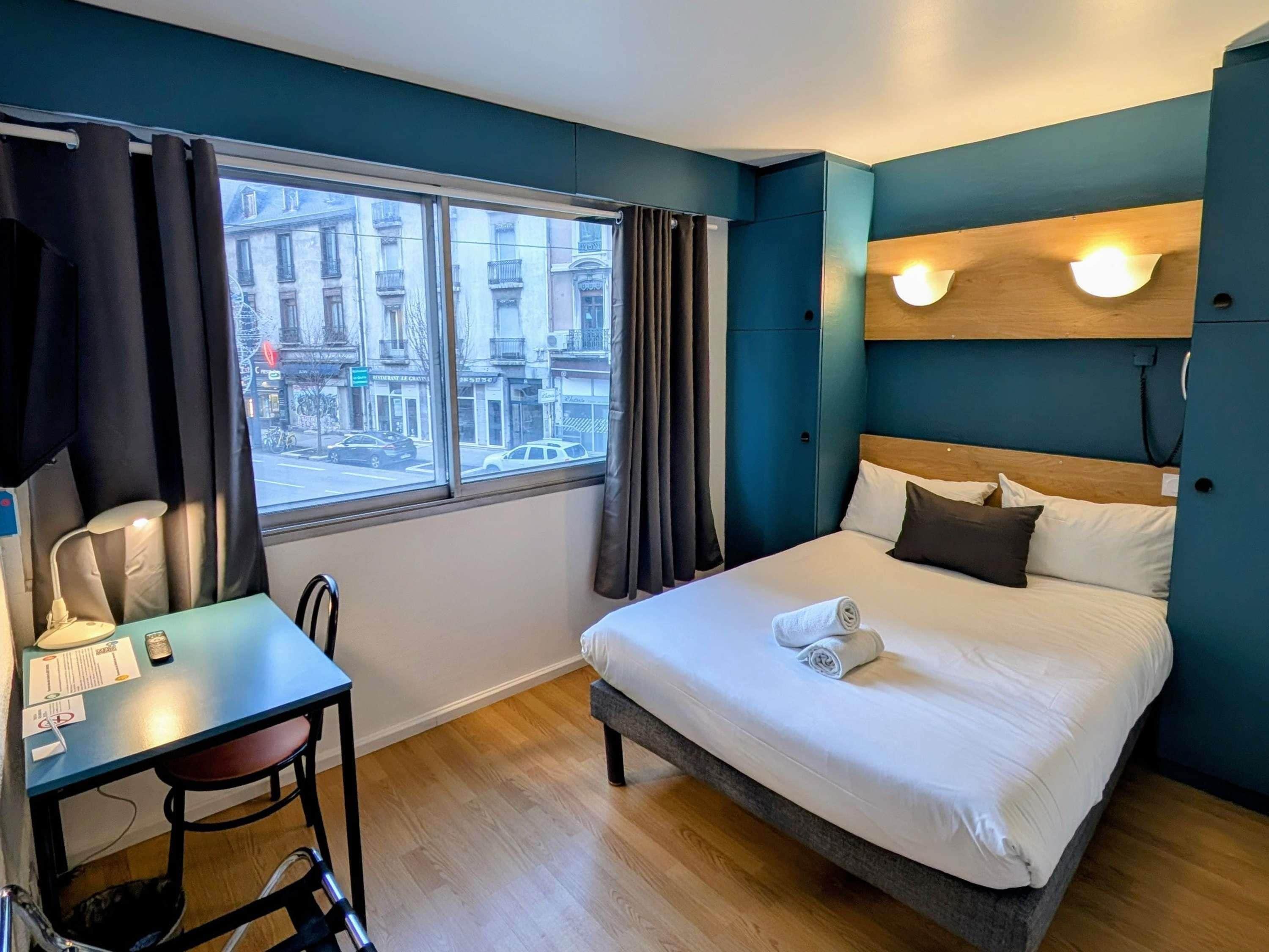 Habitación de hotel con cama, escritorio y silla. Ventana con cortinas y vistas a edificios.