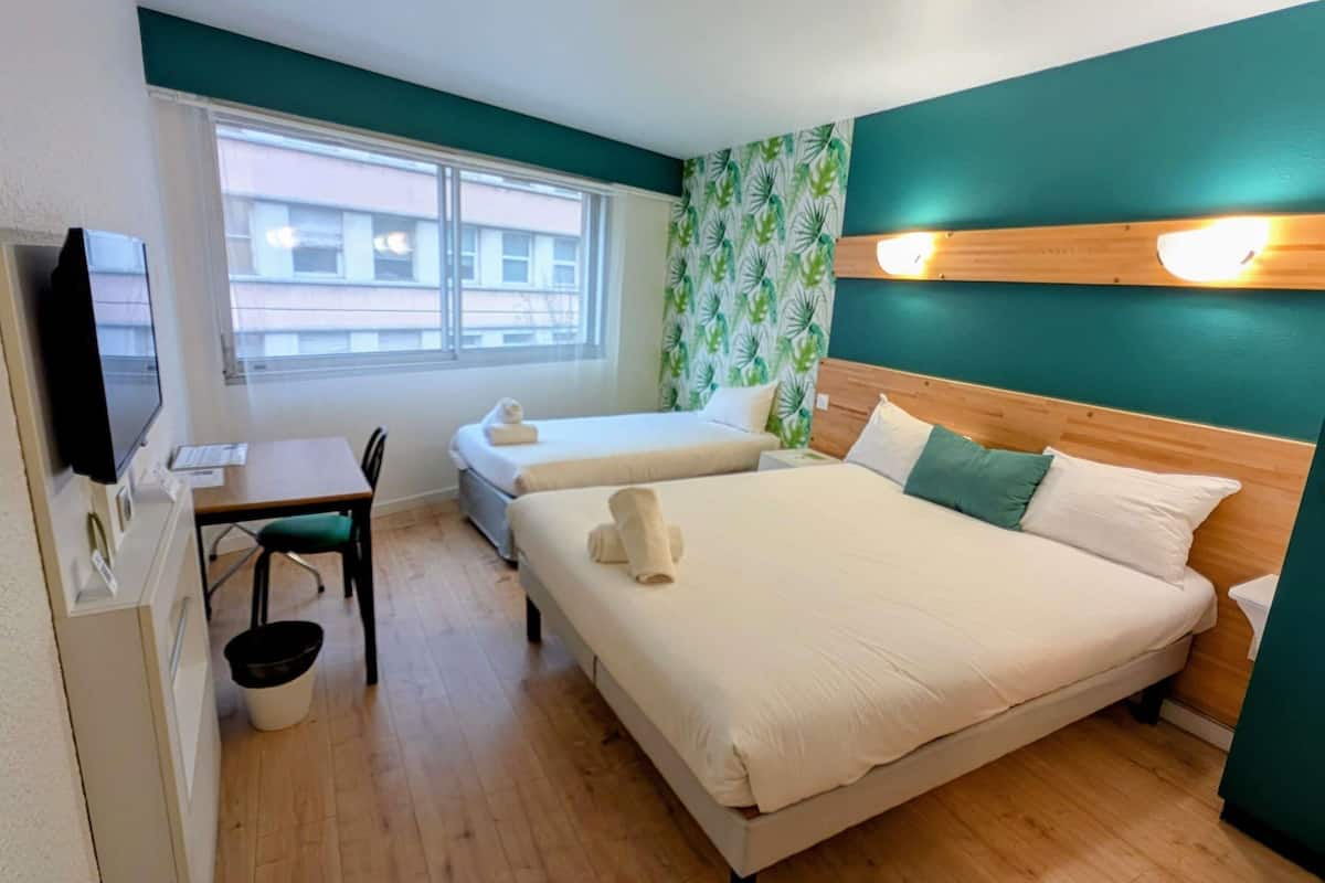 A hotel room with two beds, a desk, a chair, a television, and a window with a view of buildings.