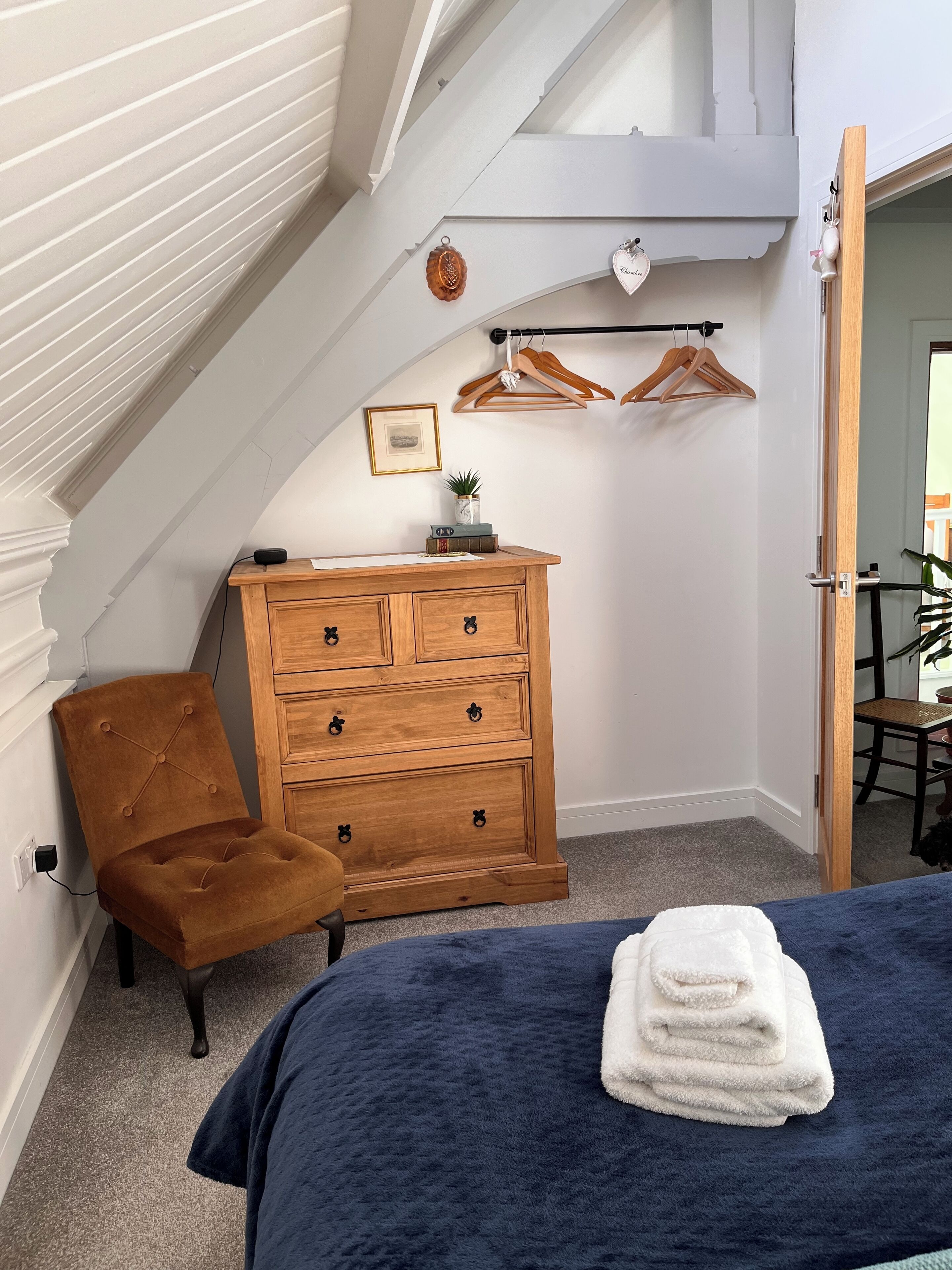 A bedroom with a bed, a wooden dresser, a brown chair, a coat rack with hangers, and a small plant on the dresser.
