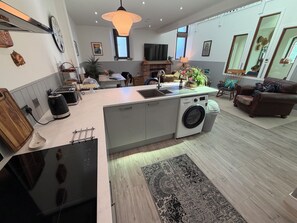 A modern kitchen with a built-in washing machine, a dining area with a table and chairs, and a living room with a sofa and decorative items. - 1902 chapel. Family friendly. Parking & garden.  (Sandown)