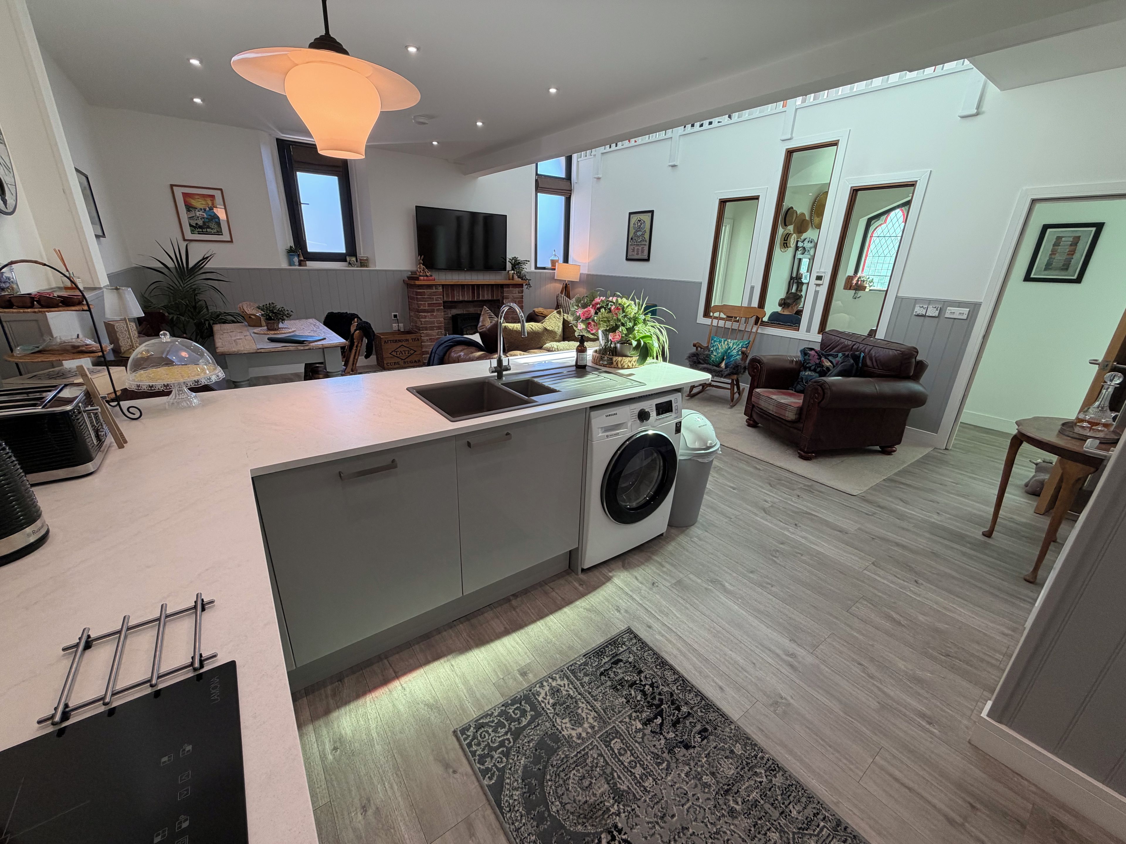Une cuisine moderne équipée d’un lave-linge intégré, un coin repas avec une table et des chaises, et un salon avec un canapé et une grande fenêtre.
