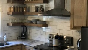 Une cuisine avec des murs recouverts de carreaux blancs, des étagères en bois où sont posés des bocaux et des assiettes, un poêle noir et une machine à café.