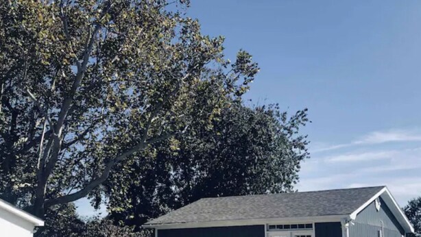 Une petite maison à un seul étage, à l’extérieur sombre, avec un porche à l’avant doté de deux chaises et un grand arbre en arrière-plan.