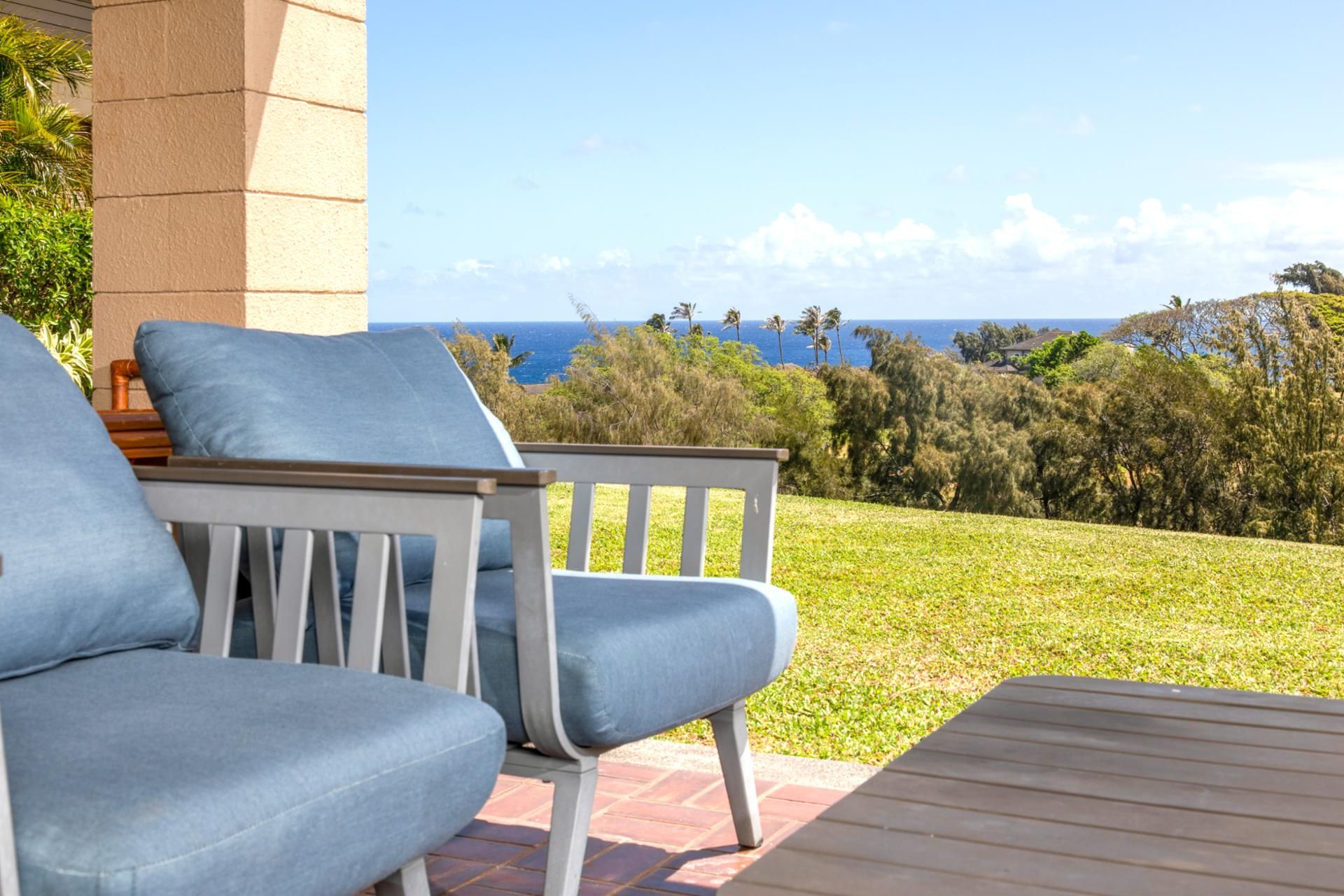Un patio con dos sillas azules acolchadas y una mesa de madera, con vistas a una zona verde con árboles y el océano.