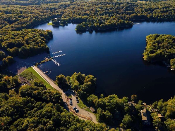 Une vue aérienne d’un lac entouré d’une forêt dense, avec un quai et une aire de stationnement visibles.