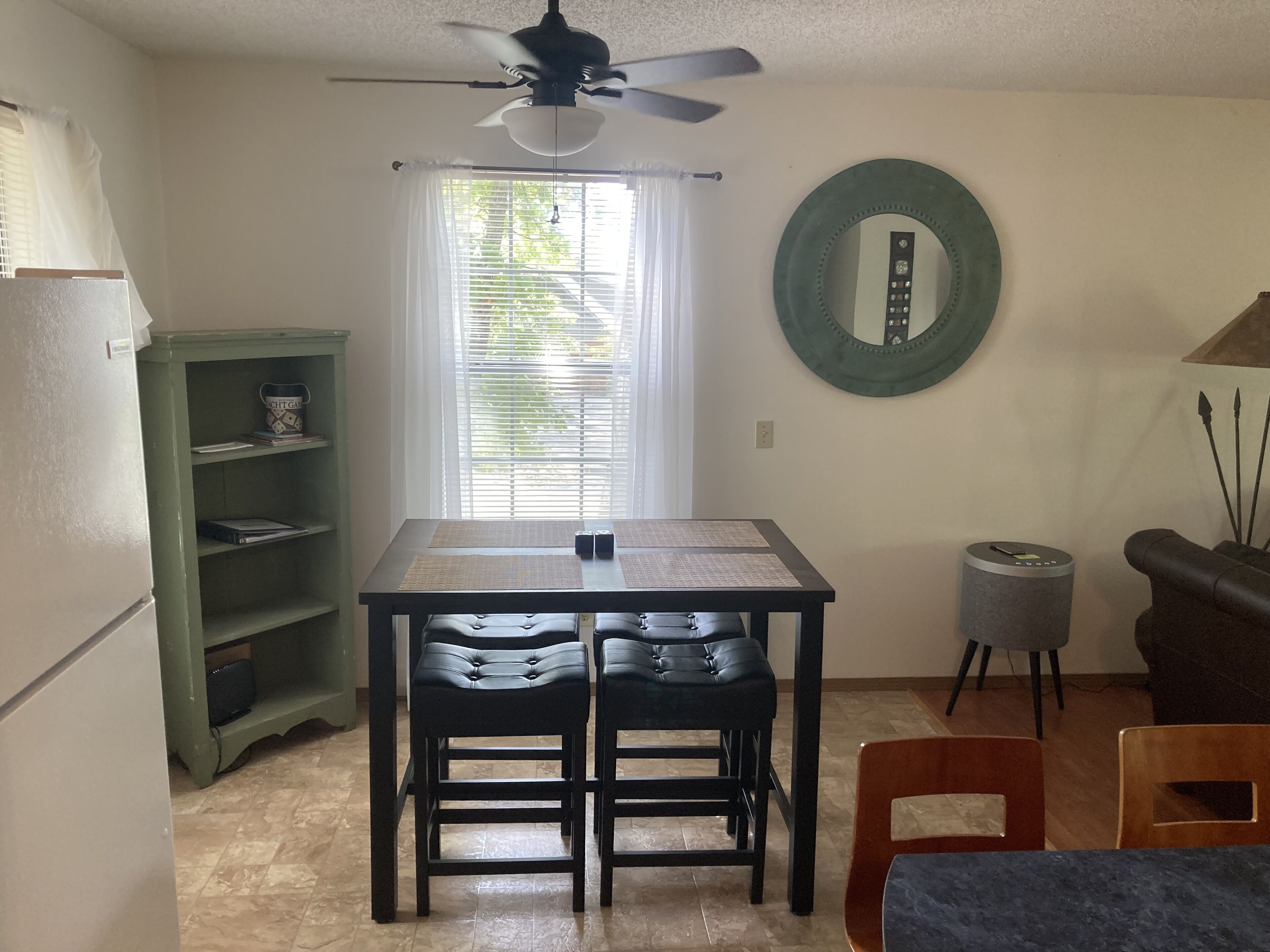 A compact kitchen with a dining table, chairs, a ceiling fan, a window with blinds, a green shelf, a round mirror, and a small stool.