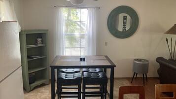 A compact kitchen with a dining table, chairs, a ceiling fan, a window with blinds, a green shelf, a round mirror, and a small stool.