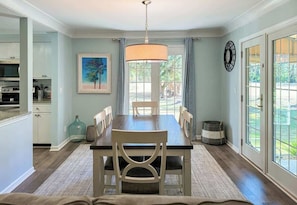 A dining area with a wooden table, chairs, a hanging light fixture, a painting on the wall, and a view through the glass door.