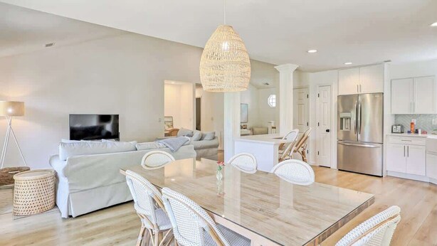 A modern living room with a sofa, dining table, wicker chairs, a TV, and a kitchen area with a refrigerator.