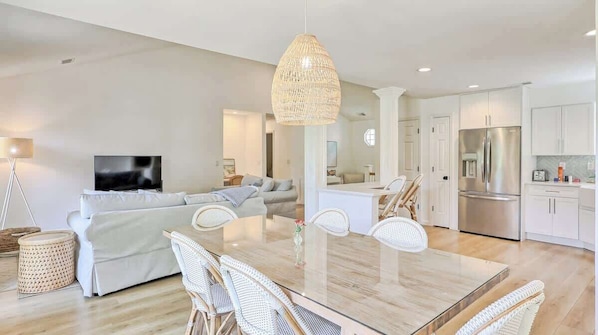 A modern living room with a sofa, dining table, wicker chairs, a TV, and a kitchen area with a refrigerator.